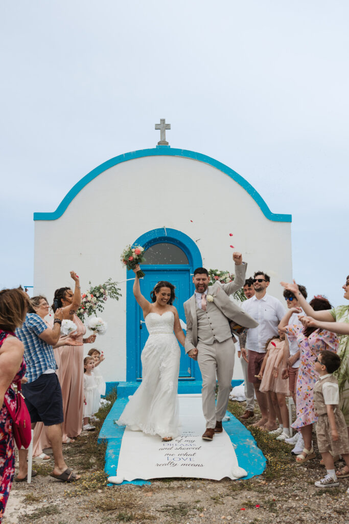 Outdoors island wedding ceremony, editorial photography, destination wedding at Agios Stephanos, Kastri island in KOS, Greece. Confetti photo with rice being thrown. Blue and white chapel.