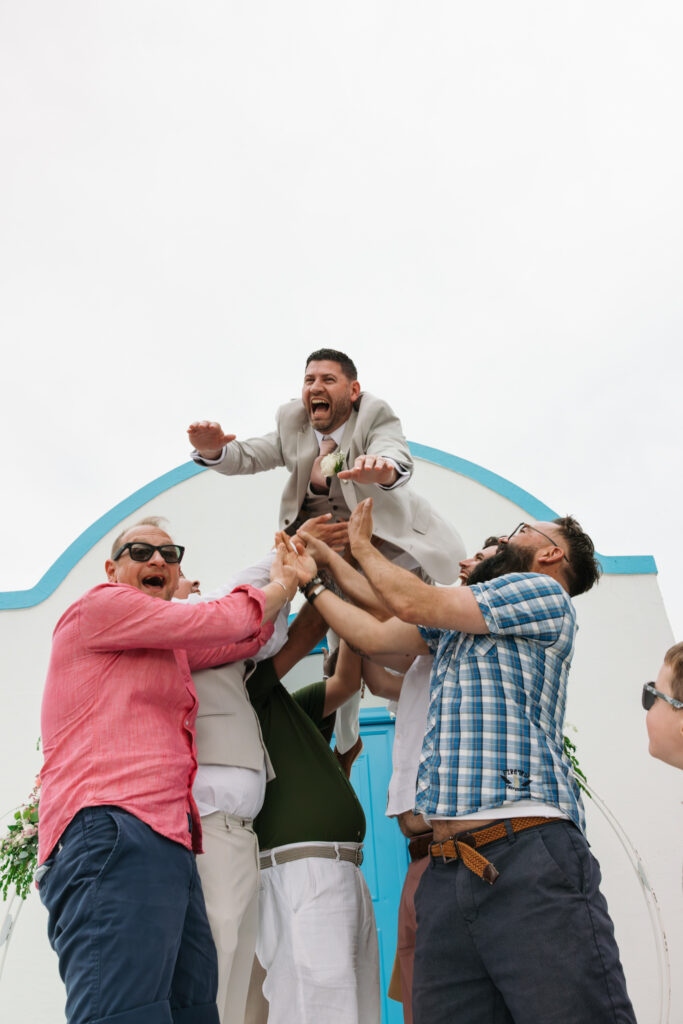 Outdoors island wedding ceremony, editorial photography, destination wedding at Agios Stephanos, Kastri island in KOS, Greece. Blue and white chapel. Groom thrown in the air by groomsmen.