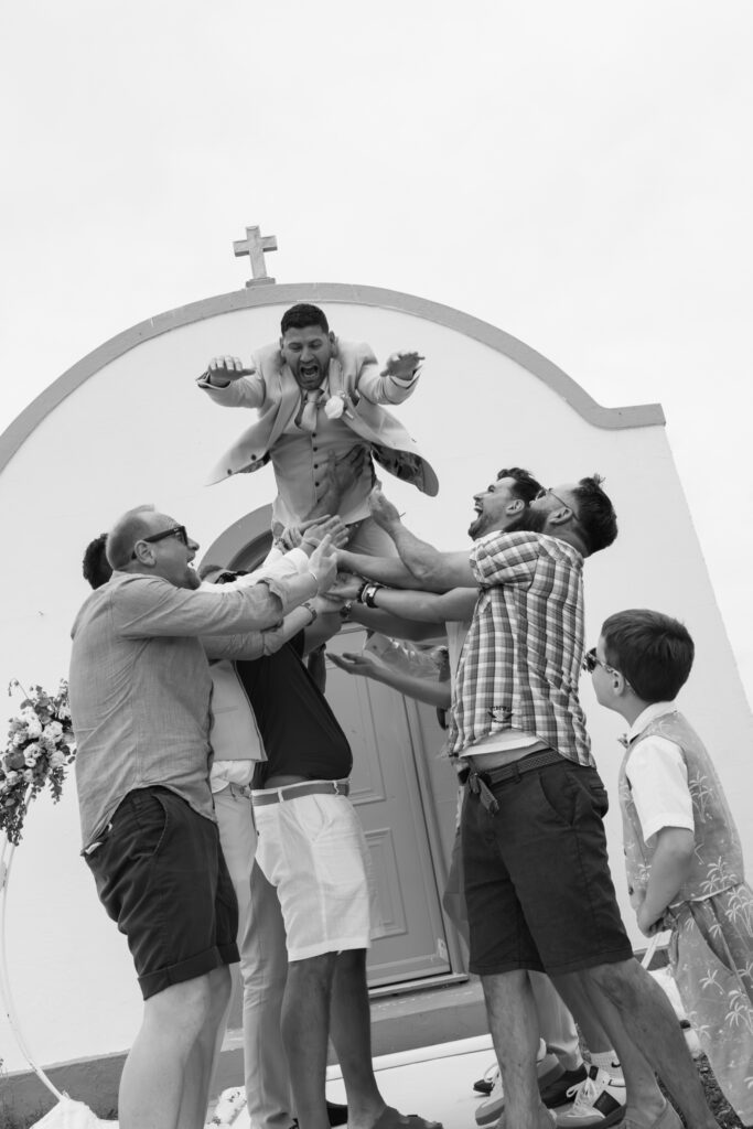 Outdoors island wedding ceremony, editorial photography, destination wedding at Agios Stephanos, Kastri island in KOS, Greece. Blue and white chapel. Groom thrown in the air by groomsmen.