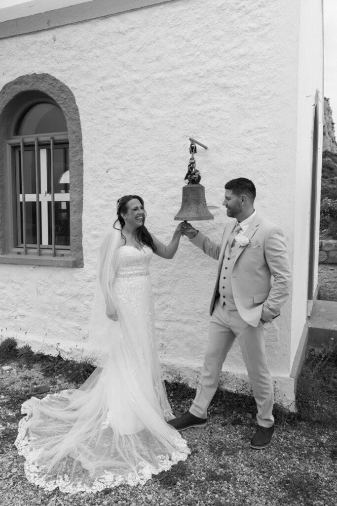 Outdoors island wedding ceremony, editorial photography, destination wedding at Agios Stephanos, Kastri island in KOS, Greece. Blue and white chapel. Bride and groom ringing the bell.