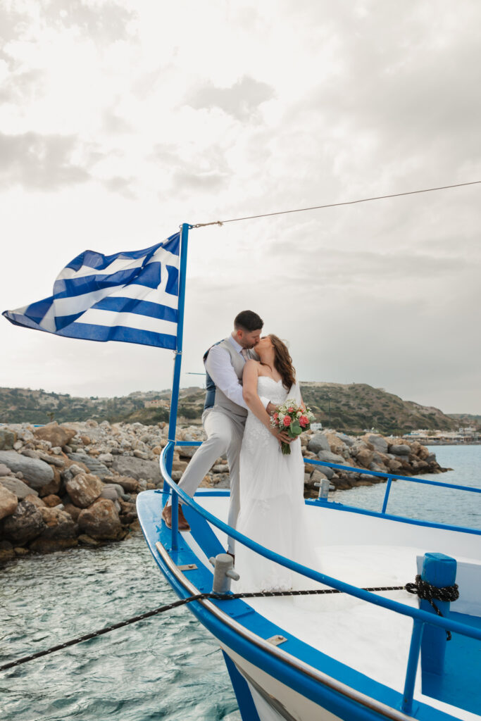 Outdoors island wedding ceremony, editorial photography, destination wedding at Agios Stephanos, Kastri island in KOS, Greece. Couple portraits on the boat that took the wedding guests from the island to the beach, with a greek flag.