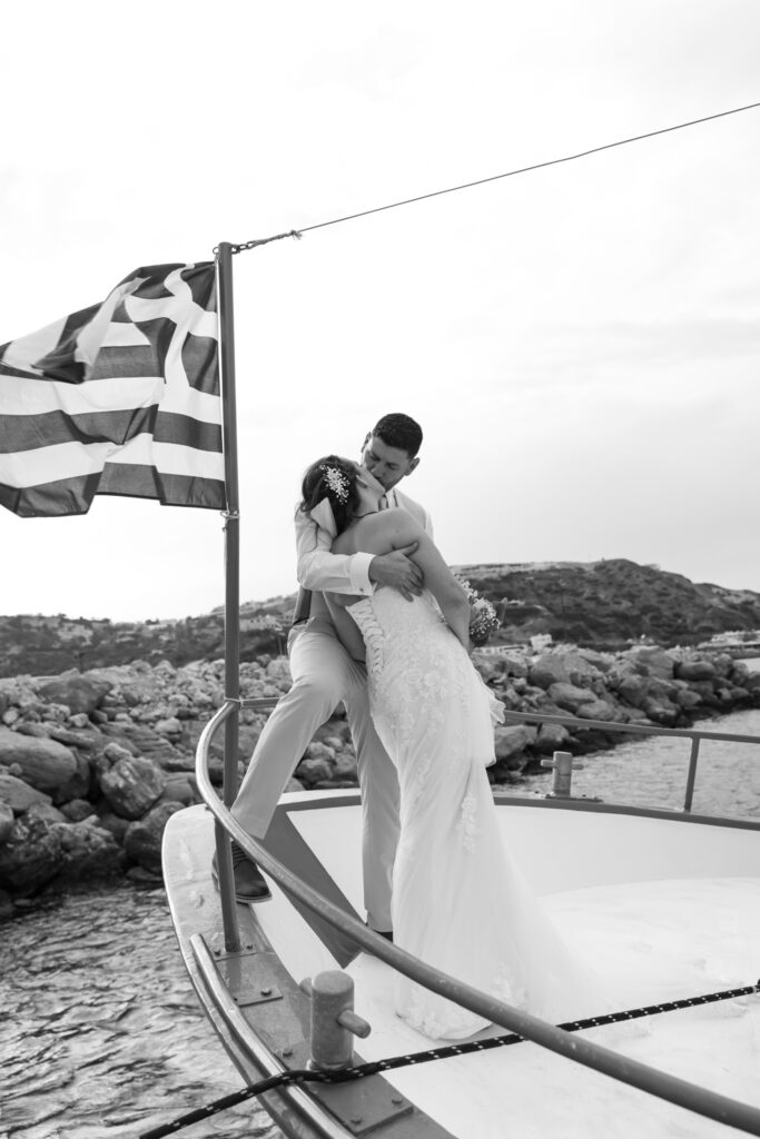 Outdoors island wedding ceremony, editorial photography, destination wedding at Agios Stephanos, Kastri island in KOS, Greece. Couple portraits on the boat that took the wedding guests from the island to the beach, with a greek flag.