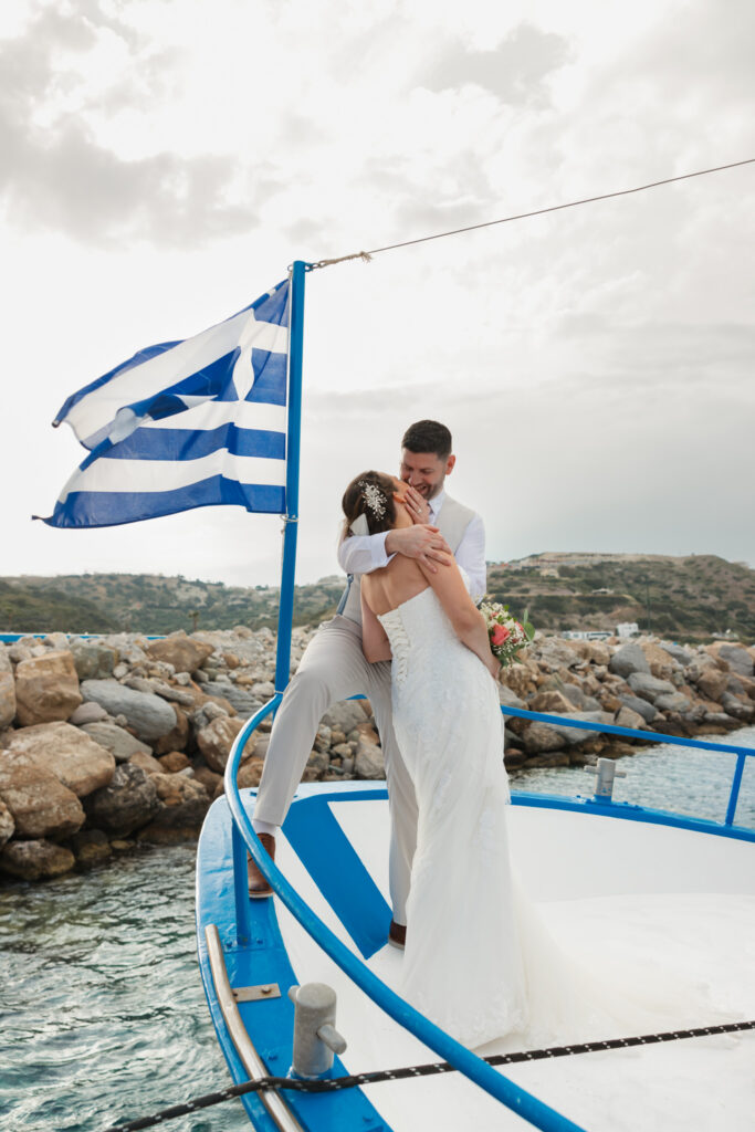 Outdoors island wedding ceremony, editorial photography, destination wedding at Agios Stephanos, Kastri island in KOS, Greece. Couple portraits on the boat that took the wedding guests from the island to the beach, with a greek flag.