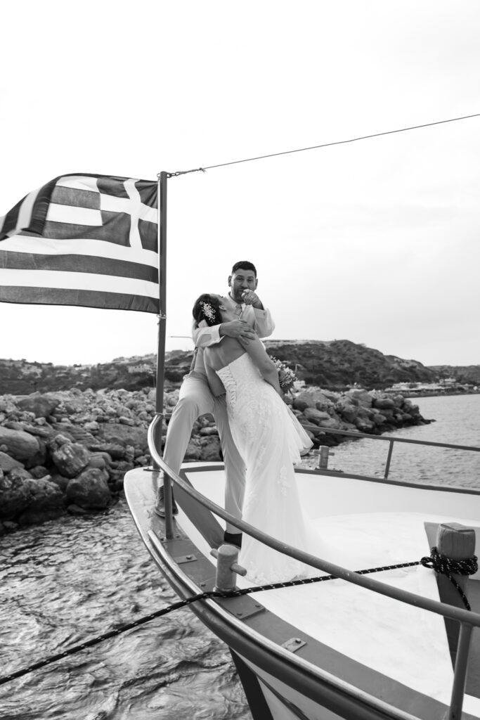 Outdoors island wedding ceremony, editorial photography, destination wedding at Agios Stephanos, Kastri island in KOS, Greece. Couple portraits on the boat that took the wedding guests from the island to the beach, with a greek flag.
