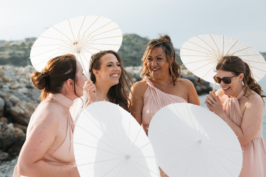 Outdoors island wedding ceremony, editorial photography, destination wedding at Agios Stephanos, Kastri island in KOS, Greece. Bridesmaids with bride photos bu the pier, with paper parasols. Bridesmaids wearing baby pink multi style dresses.