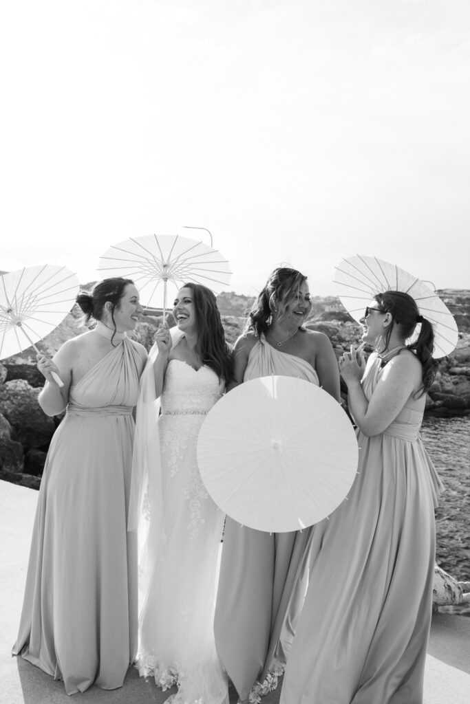 Outdoors island wedding ceremony, editorial photography, destination wedding at Agios Stephanos, Kastri island in KOS, Greece. Bridesmaids with bride photos bu the pier, with paper parasols. Bridesmaids wearing baby pink multi style dresses.