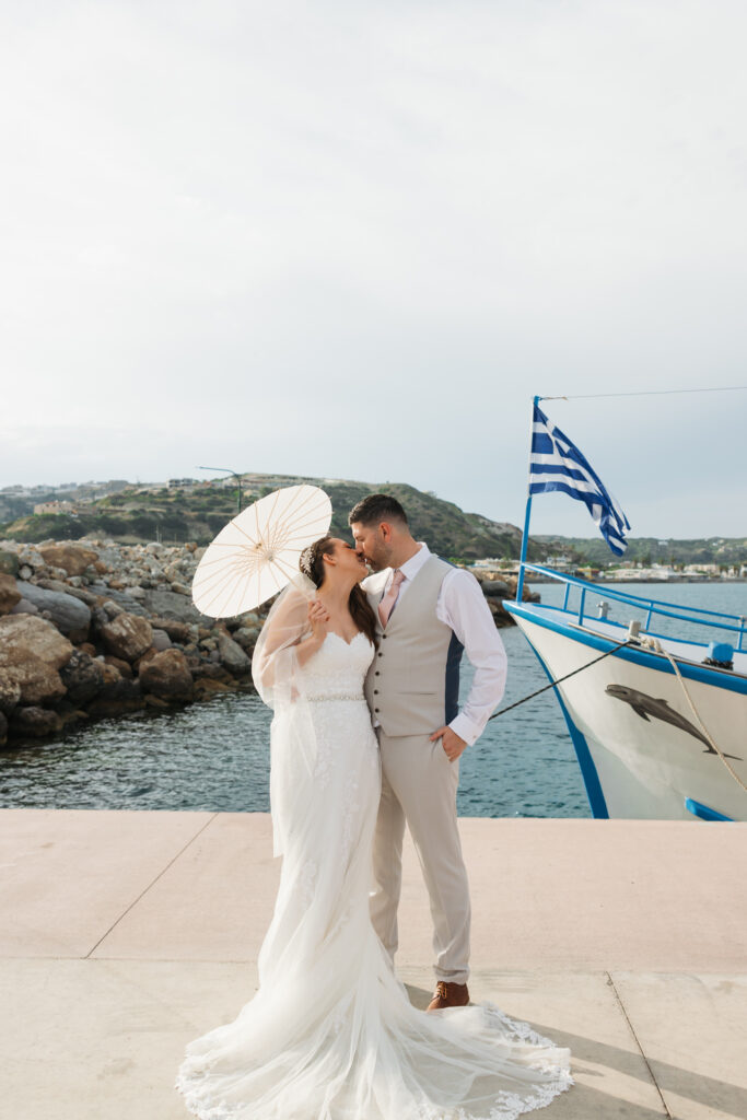 Outdoors island wedding ceremony, editorial photography, destination wedding at Agios Stephanos, Kastri island in KOS, Greece. Bride and groom couple photos by the pier, with boat, greek flag and paper parasol.