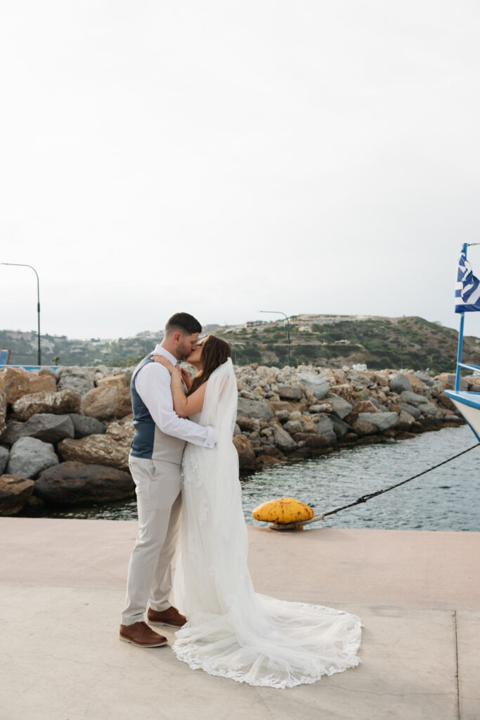 Outdoors island wedding ceremony, editorial photography, destination wedding at Agios Stephanos, Kastri island in KOS, Greece. Bride and groom couple photos by the pier, with boat, greek flag.