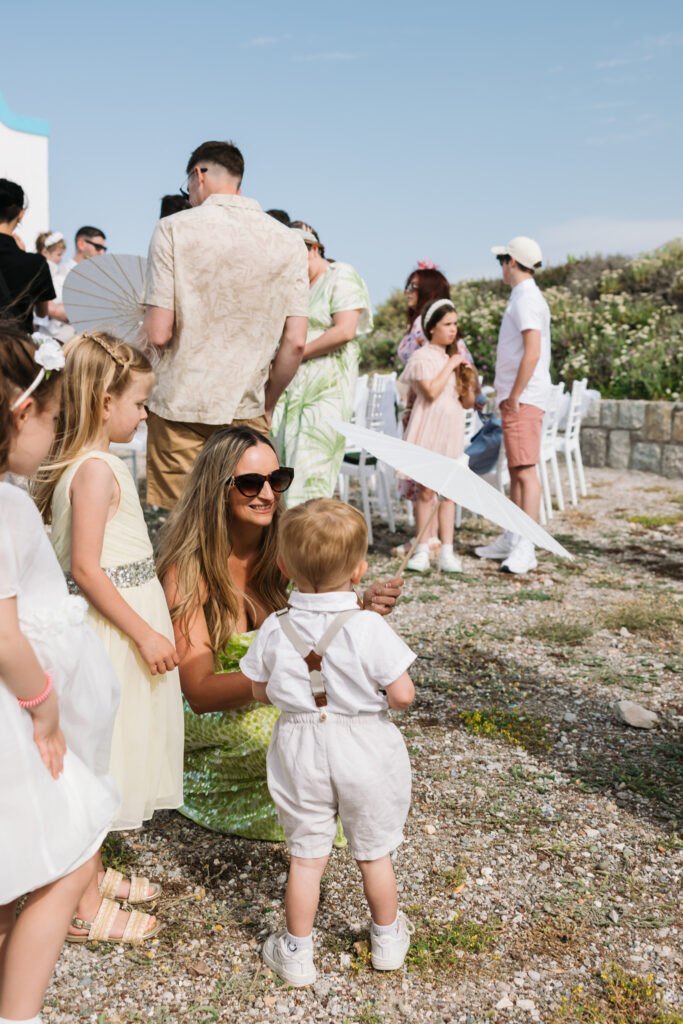 Wedding guests with paper parasols at outdoors island wedding ceremony, editorial photography, destination wedding at Agios Stephanos, Kastri island in KOS, Greece.