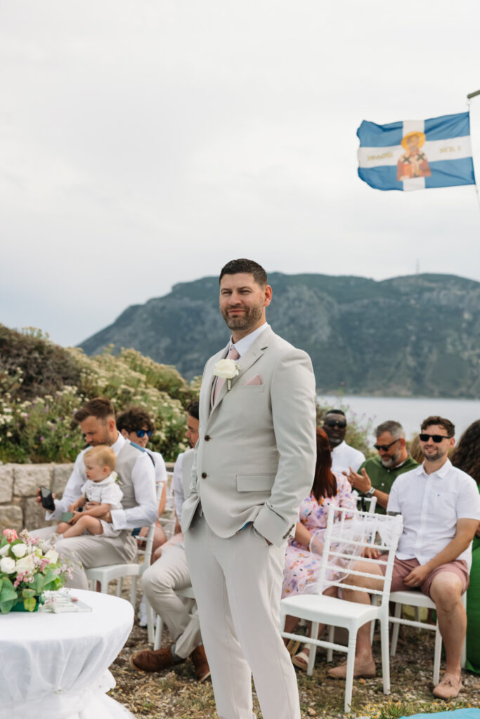 Groom waiting at the alter for his bride, editorial photography, destination wedding at Agios Stephanos, Kastri island in KOS, Greece. Groom wearing light beige suit and baby pink tie.