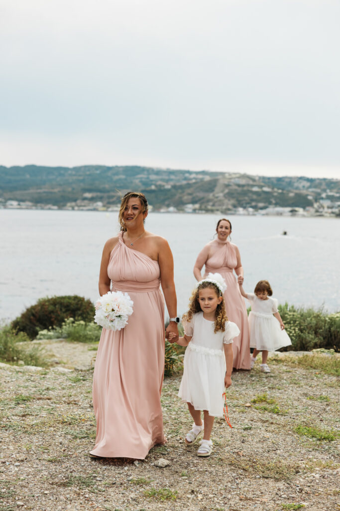 Bridesmaids walking down the aisle with her bridesmaid, editorial photography, destination wedding at Agios Stephanos, Kastri island in KOS, Greece. Bridesmaid wearing pink dress.