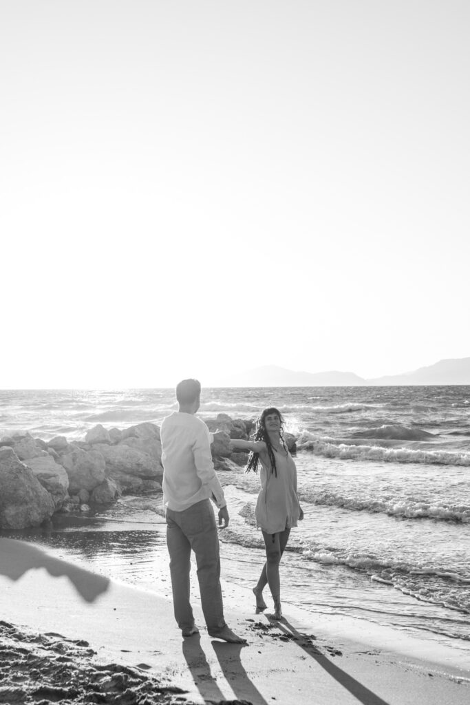 Romantic storytelling portrait of Greek and Italian couple walking barefoot on Kos beach at golden hour — destination wedding photographer Greece. Editorial photography, engagement beach sunset session.
