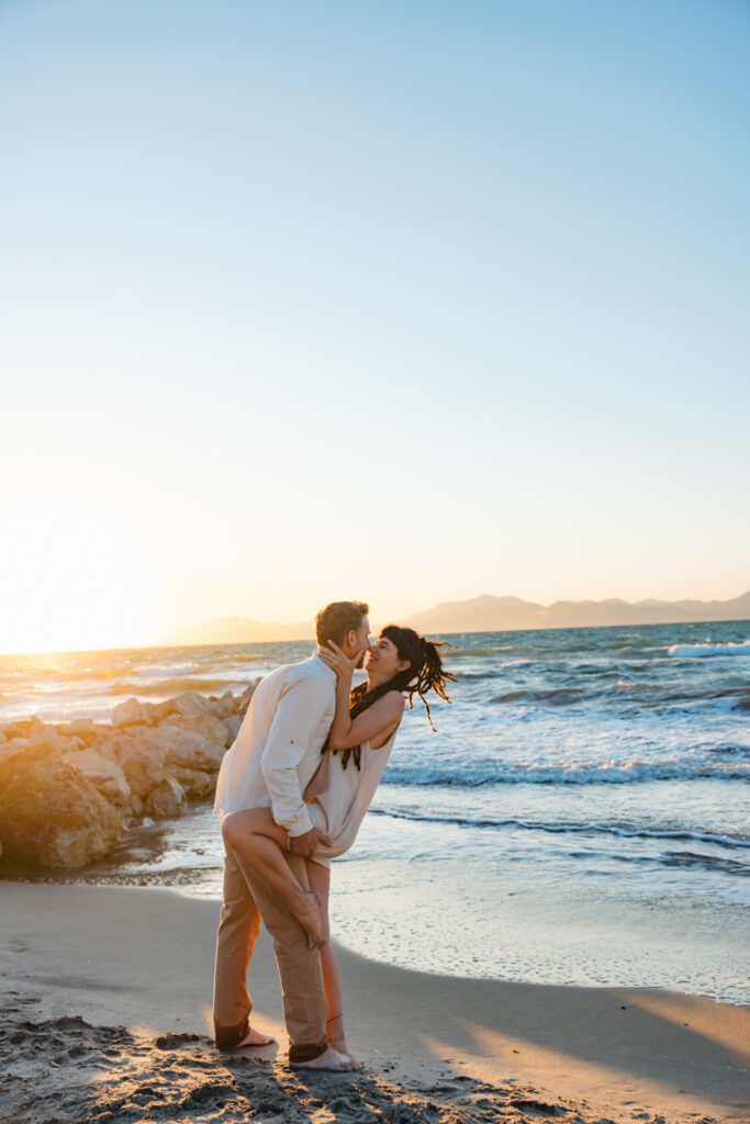 Romantic storytelling portrait of Greek and Italian couple walking barefoot on Kos beach at golden hour — destination wedding photographer Greece. Editorial photography, engagement beach sunset session.