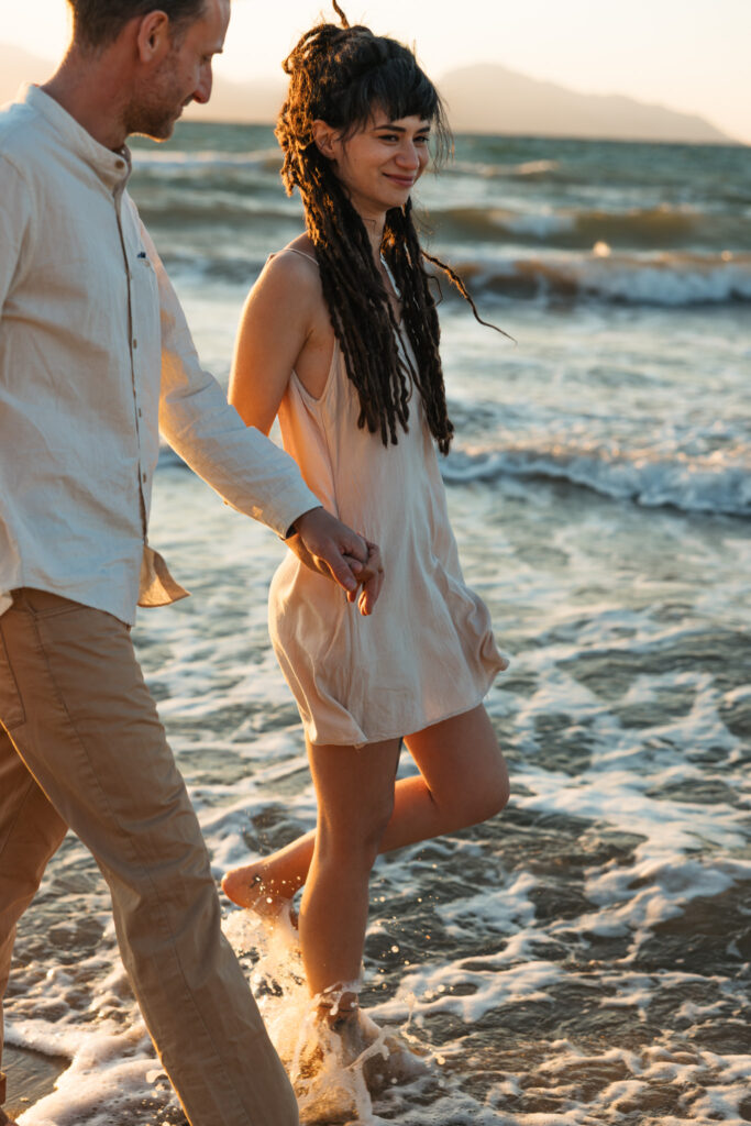 Romantic storytelling portrait of Greek and Italian couple walking barefoot on Kos beach at golden hour — destination wedding photographer Greece. Editorial photography, engagement beach sunset session.