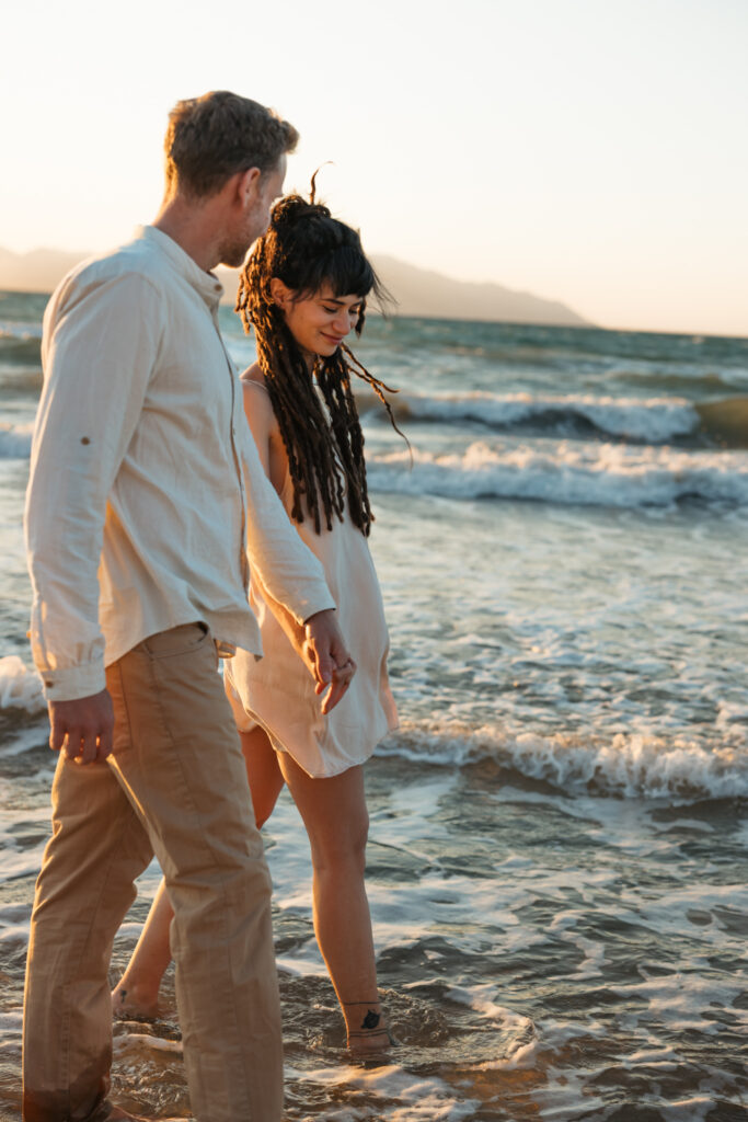 Romantic storytelling portrait of Greek and Italian couple walking barefoot on Kos beach at golden hour — destination wedding photographer Greece. Editorial photography, engagement beach sunset session.