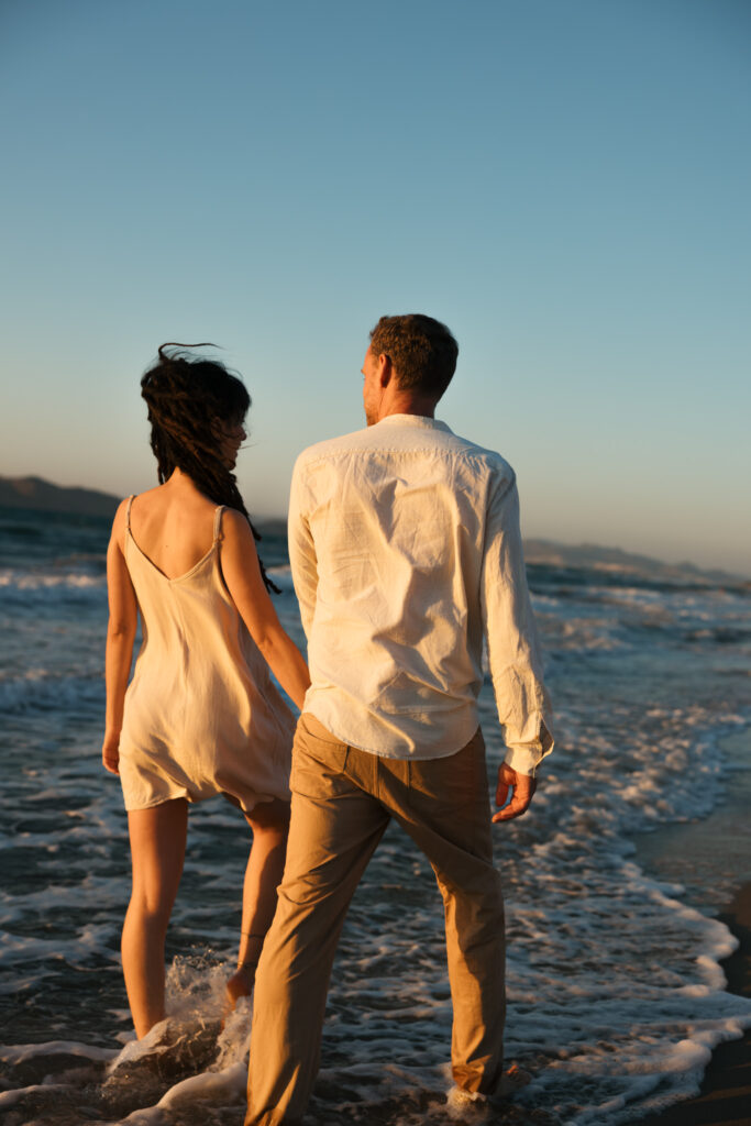 Romantic storytelling portrait of Greek and Italian couple walking barefoot on Kos beach at golden hour — destination wedding photographer Greece. Editorial photography, engagement beach sunset session.