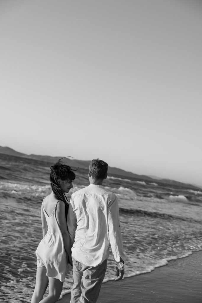 Romantic storytelling portrait of Greek and Italian couple walking barefoot on Kos beach at golden hour — destination wedding photographer Greece. Editorial photography, engagement beach sunset session.