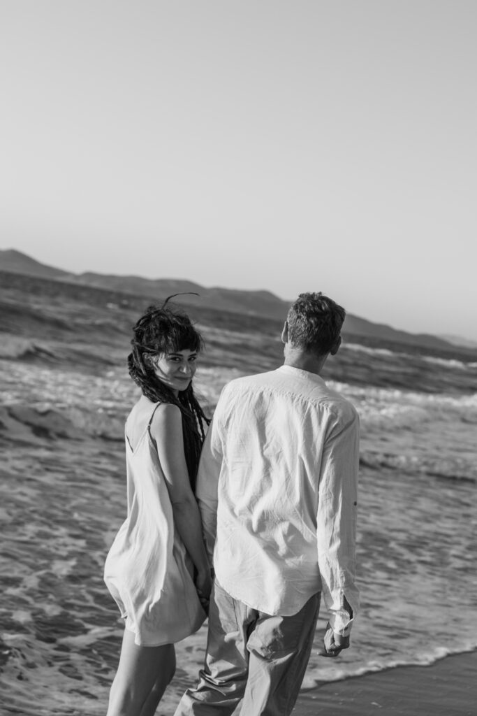 Romantic storytelling portrait of Greek and Italian couple walking barefoot on Kos beach at golden hour — destination wedding photographer Greece. Editorial photography, engagement beach sunset session.