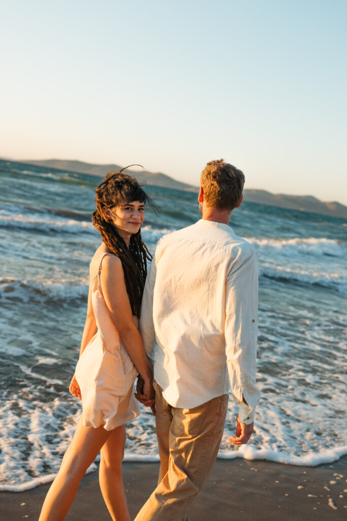 Romantic storytelling portrait of Greek and Italian couple walking barefoot on Kos beach at golden hour — destination wedding photographer Greece. Editorial photography, engagement beach sunset session.