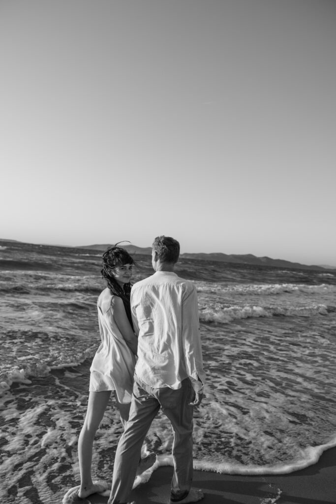 Romantic storytelling portrait of Greek and Italian couple walking barefoot on Kos beach at golden hour — destination wedding photographer Greece. Editorial photography, engagement beach sunset session.