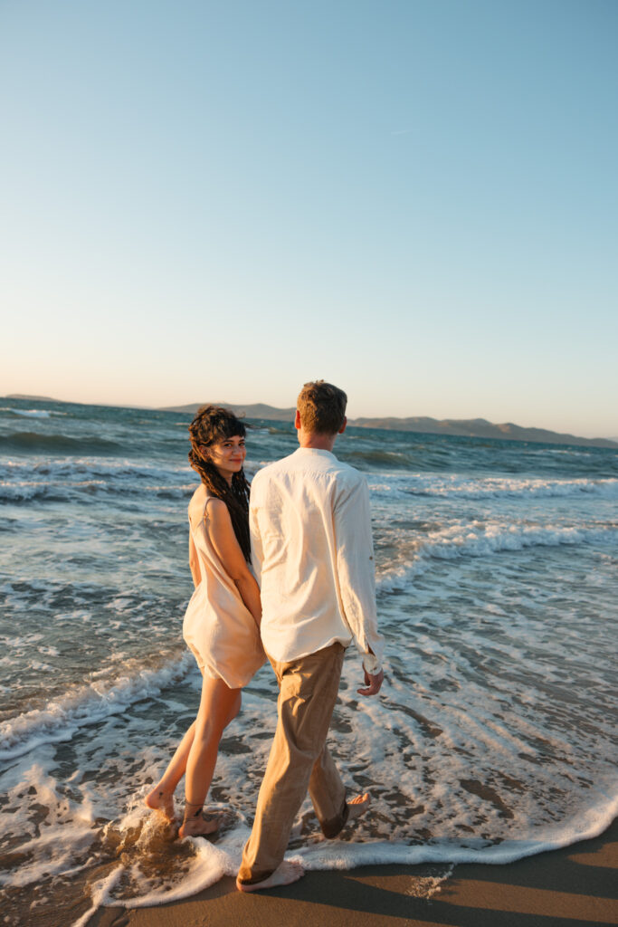 Romantic storytelling portrait of Greek and Italian couple walking barefoot on Kos beach at golden hour — destination wedding photographer Greece. Editorial photography, engagement beach sunset session.