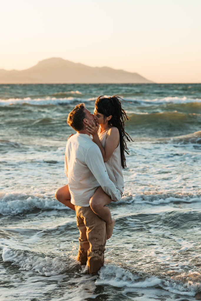 Romantic storytelling portrait of Greek and Italian couple walking barefoot on Kos beach at golden hour — destination wedding photographer Greece. Editorial photography, engagement beach sunset session.