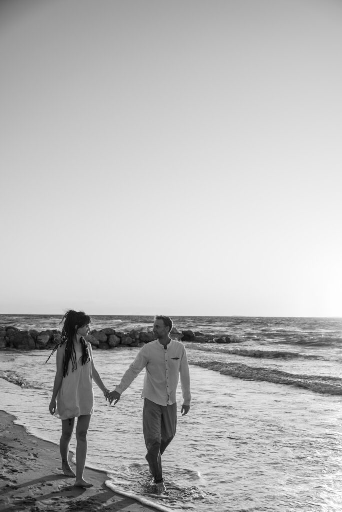 Romantic storytelling portrait of Greek and Italian couple walking barefoot on Kos beach at golden hour — destination wedding photographer Greece. Editorial photography, engagement beach sunset session.