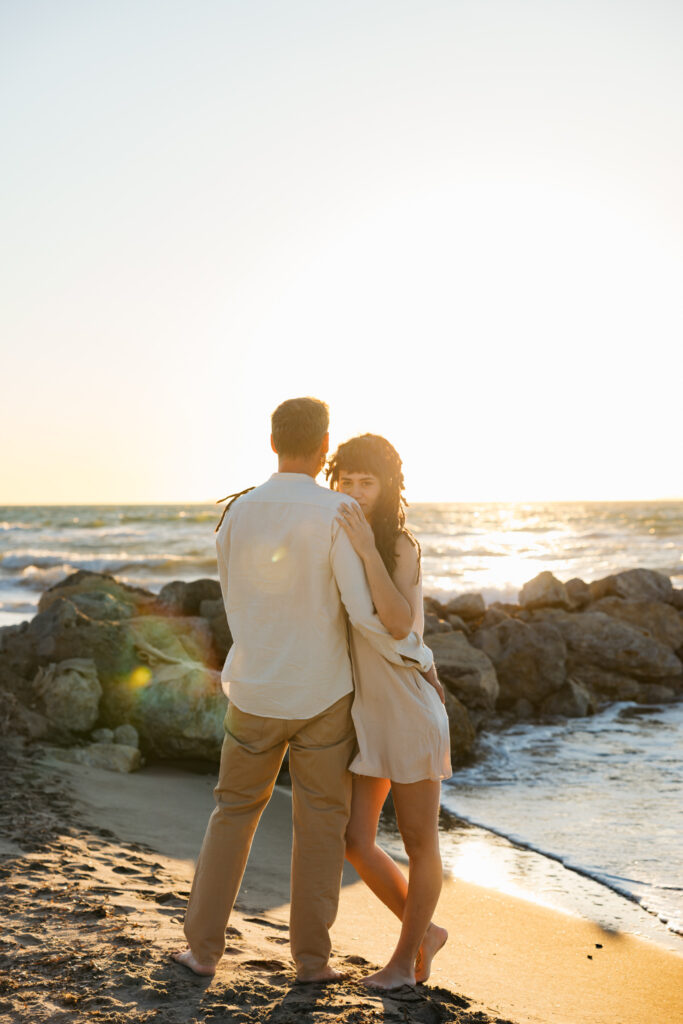 Romantic storytelling portrait of Greek and Italian couple walking barefoot on Kos beach at golden hour — destination wedding photographer Greece. Editorial photography, engagement beach sunset session.