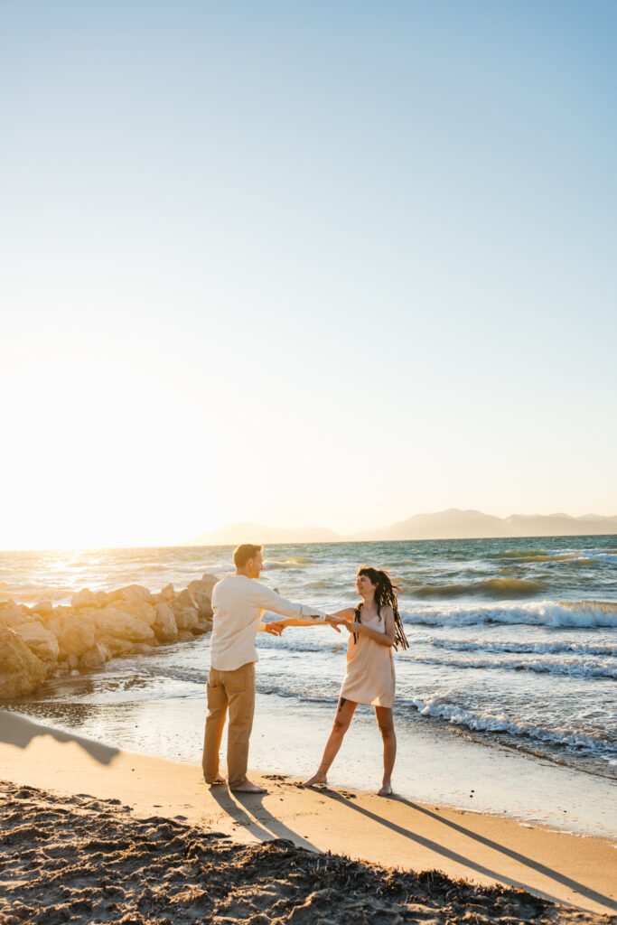 Romantic storytelling portrait of Greek and Italian couple walking barefoot on Kos beach at golden hour — destination wedding photographer Greece. Editorial photography, engagement beach sunset session.